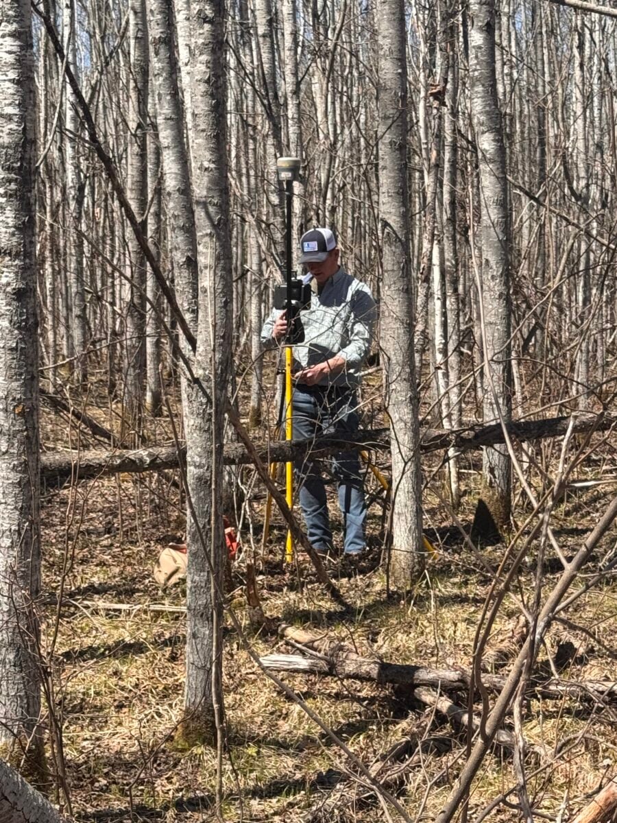 A person wearing a baseball cap uses surveying equipment on a tall yellow tripod in a forest with leafless trees and scattered undergrowth.