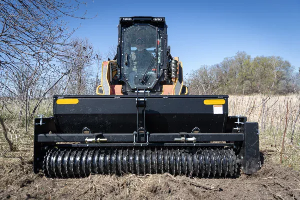 A front-facing skid steer loader equipped with a heavy-duty roller attachment is working on uneven soil, demonstrating TLM (transformation land management) in an outdoor field with trees and dry grass under a clear blue sky.