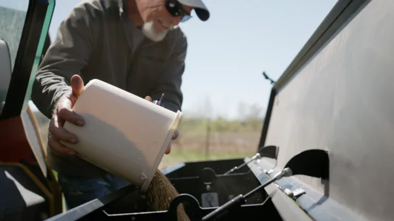 A man wearing sunglasses and a cap pours grain from a white bucket into the hopper of a machine outdoors on a sunny day, showcasing transformation land management (TLM) amid blurred fields and trees in the background.