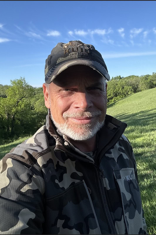 A man with a white beard wearing a camouflage jacket and cap stands outdoors in a grassy field managed by transformation land management (TLM), smiling at the camera. Trees and a blue sky with wispy clouds are visible in the background.