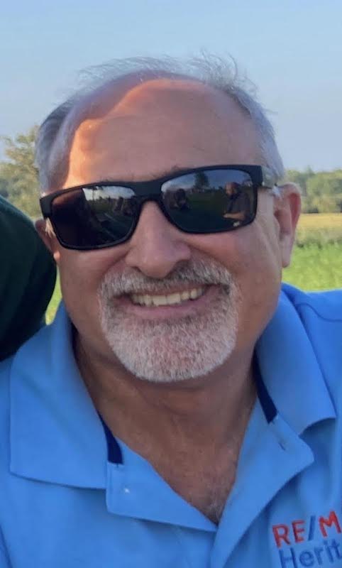 A middle-aged man with gray hair, a short white beard, and sunglasses smiles outdoors. Sporting a light blue collared shirt with TLM embroidery, he stands against a backdrop of greenery and blue sky, reflecting transformation land management values.