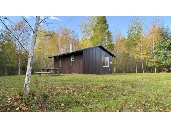 A small brown cabin with a metal roof sits on a grassy lawn surrounded by autumn trees on 80 acres near Northome MN. A wooden picnic table is placed beside the cabin under a clear sky with a few clouds.