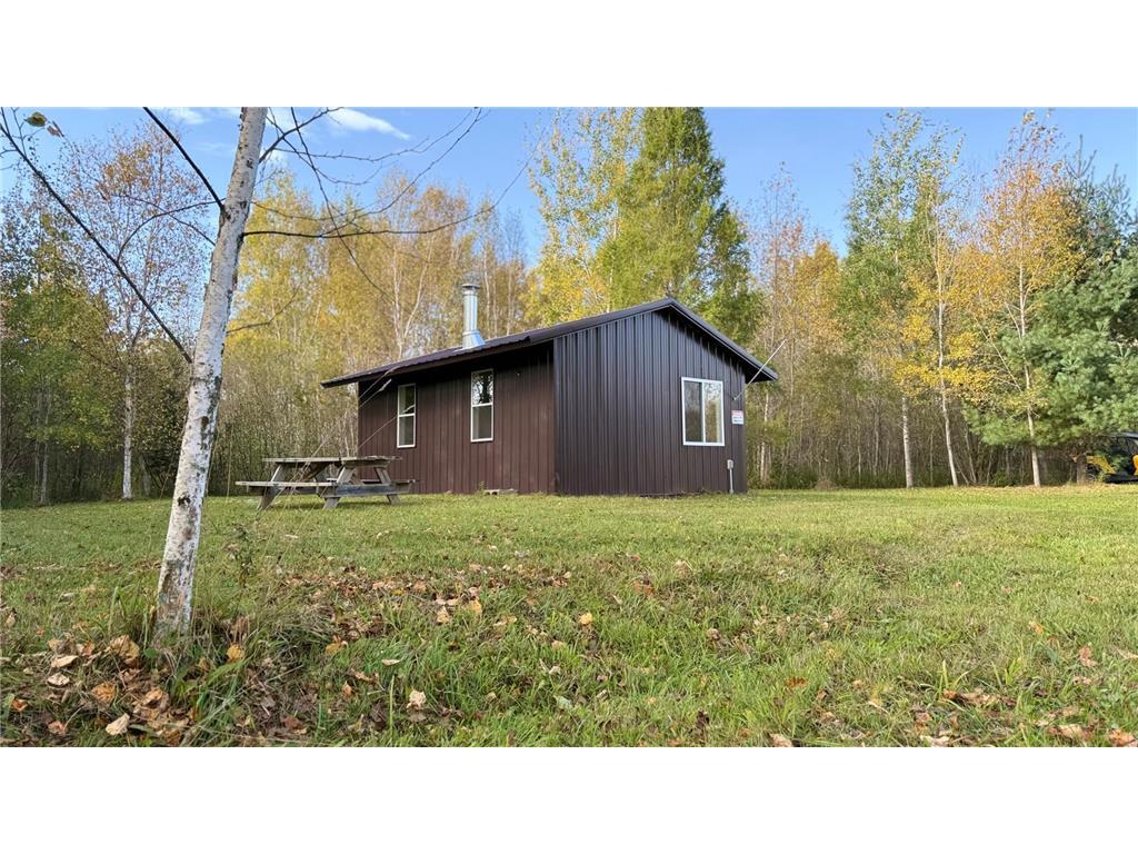 A small brown cabin with a metal roof sits on a grassy lawn surrounded by autumn trees on 80 acres near Northome MN. A wooden picnic table is placed beside the cabin under a clear sky with a few clouds.