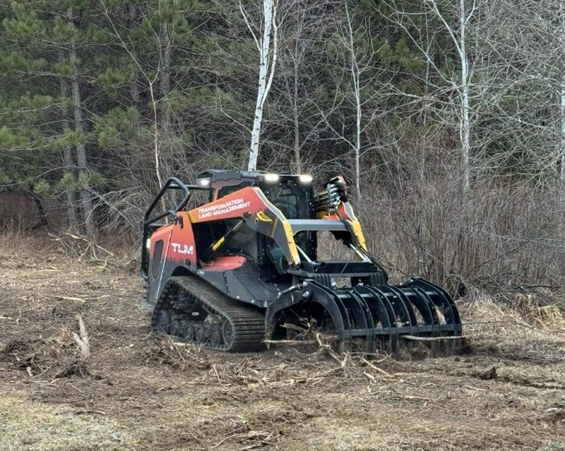A compact track loader with a grapple attachment is clearing brush and debris in a wooded area with trees and dry grass in the background.