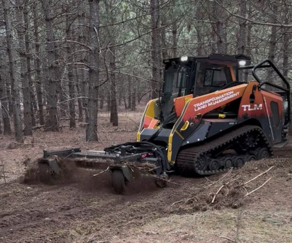 A tracked land management vehicle with “Transformation Land Management” branding clears soil and debris in a forested area, using a front-mounted attachment. Pine trees and scattered branches are visible in the background.