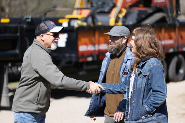 A man in a baseball cap and sunglasses shakes hands with a woman in a denim jacket, celebrating a successful TLM project, while another man stands beside her. Construction equipment and a trailer are visible in the background.