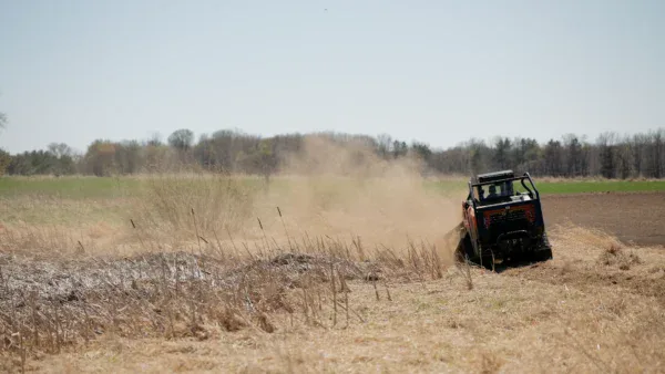A skid steer loader, part of transformation land management (TLM), moves across a dry, grassy field, kicking up a cloud of dust beneath a clear sky with distant trees lining the horizon.