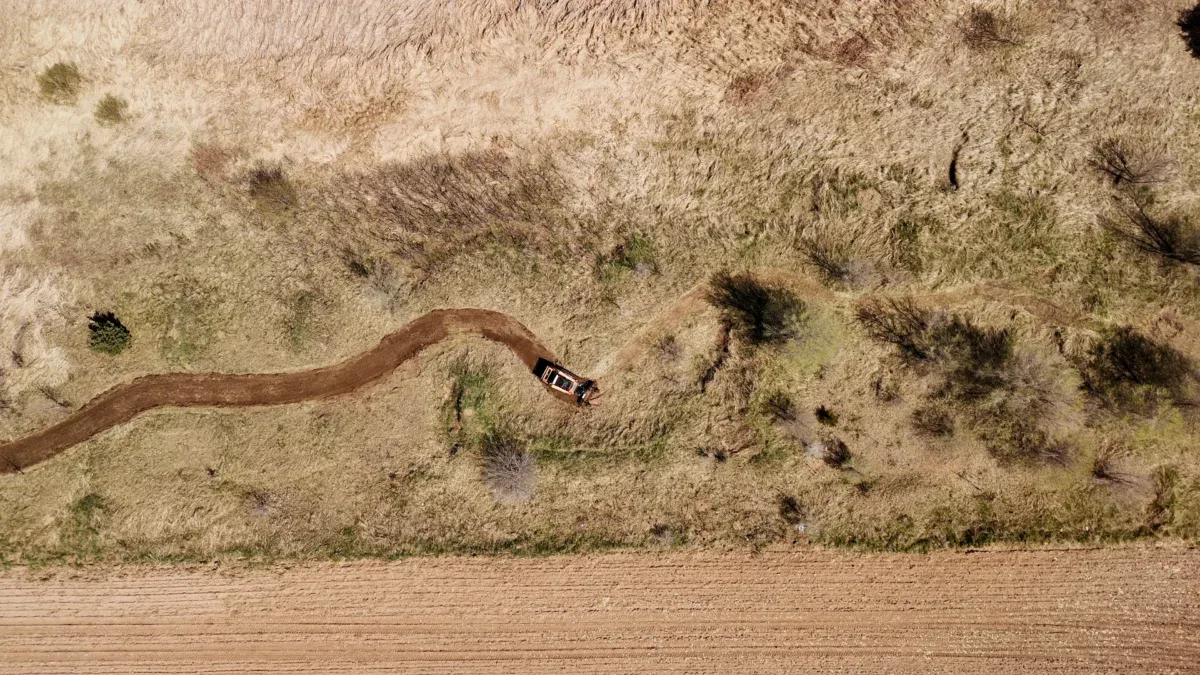 Aerial view of a dry, grassy landscape with a winding dirt path showcases transformation land management (TLM) in action. A vehicle is parked along the path, while patches of green vegetation and a bordering field hint at ongoing restoration efforts.