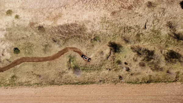 Aerial view of a dry, grassy landscape with a winding dirt path showcases transformation land management (TLM) in action. A vehicle is parked along the path, while patches of green vegetation and a bordering field hint at ongoing restoration efforts.
