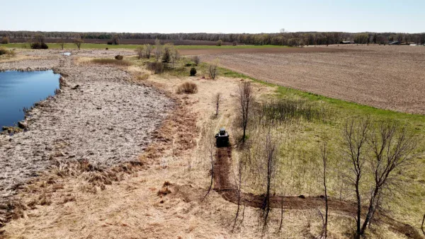 Aerial view of a rural landscape showcasing transformation land management (TLM), with a pond on the left, a curving dirt path cutting through grassy and dry areas, and a vehicle on the path. Fields and sparse trees complete the scenic background.