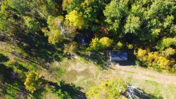 Aerial view of two small buildings surrounded by dense green and yellow trees on 160 acres near Kelliher, MN, with a dirt path and a clearing nearby. Shadows from the trees extend across the open grassy area.