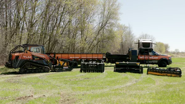 Several pieces of orange and black heavy machinery and equipment attachments for transformation land management (TLM) are arranged on a grassy field, with trees and a clear sky in the background.